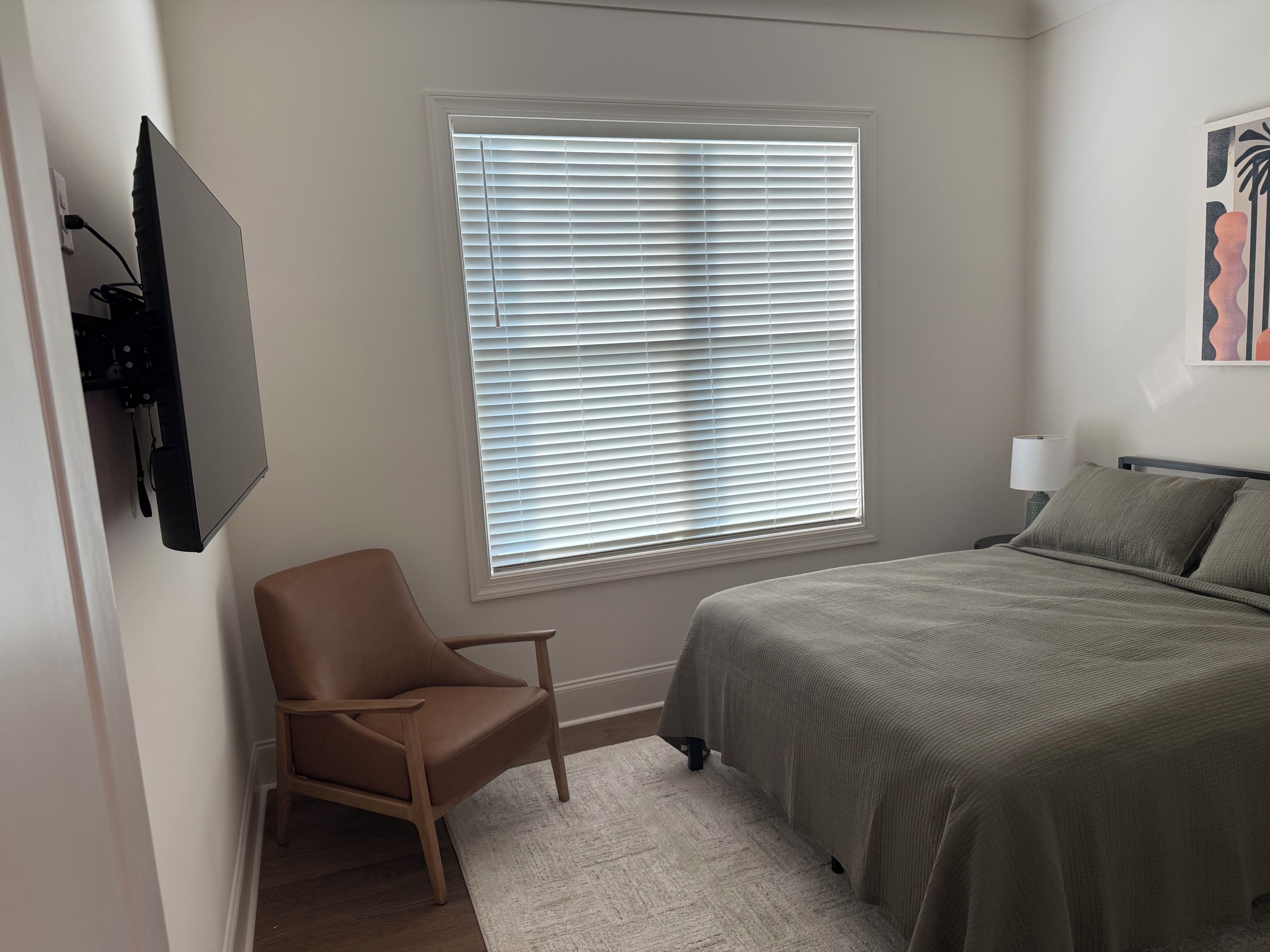 Ein Schlafzimmer mit Bett, Sessel, Fenster mit Jalousien und einem an der Wand befestigten Fernseher.