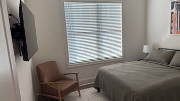 Ein Schlafzimmer mit Bett, Sessel, Fenster mit Jalousien und einem an der Wand befestigten Fernseher.