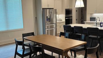 Cozinha moderna com eletrodomésticos de aço inoxidável, área de refeições com mesa de madeira e cadeiras pretas, e luminária pendente sobre a ilha.