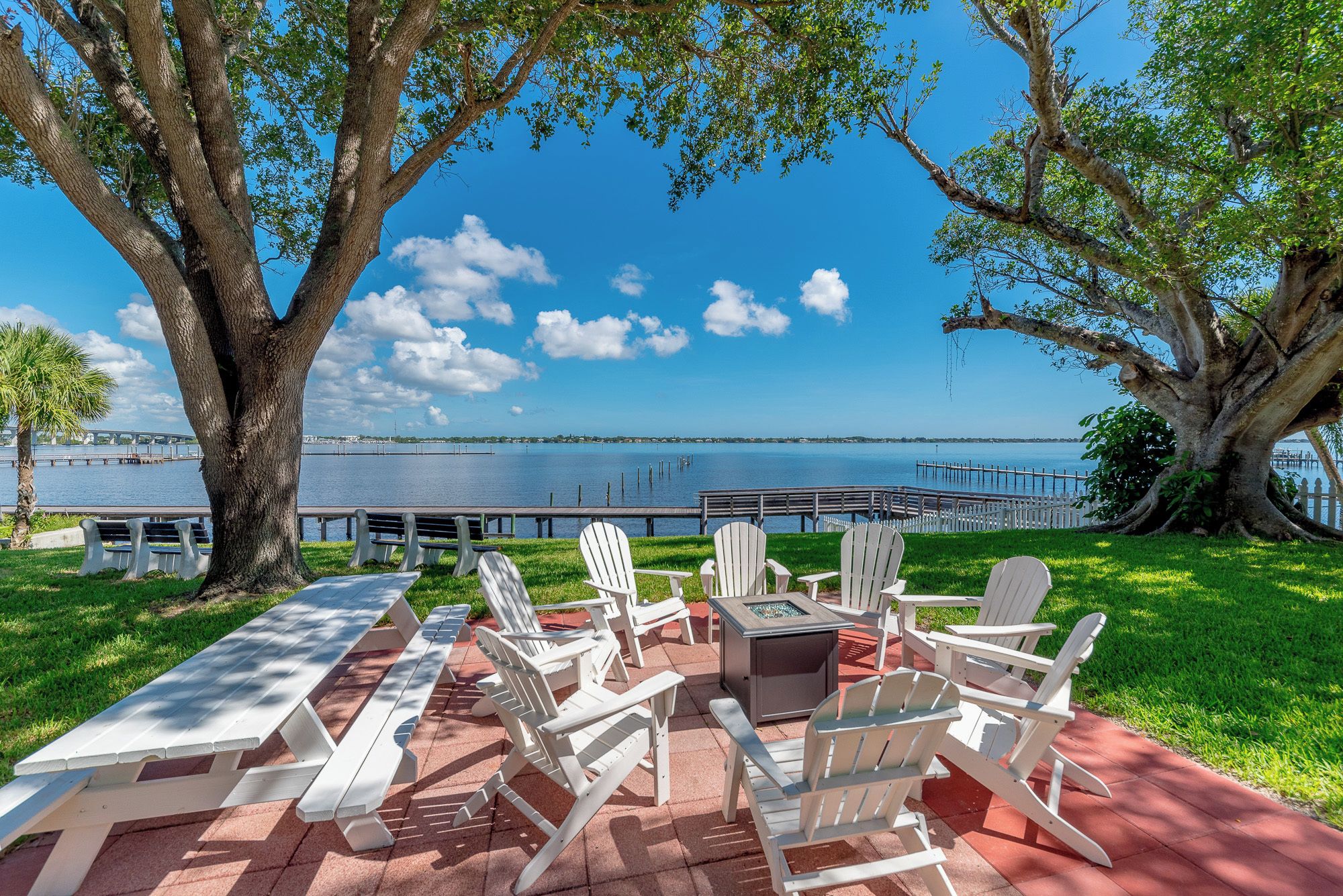A lakeside patio with Adirondack chairs, a fire pit, and a view of a bridge over the water.