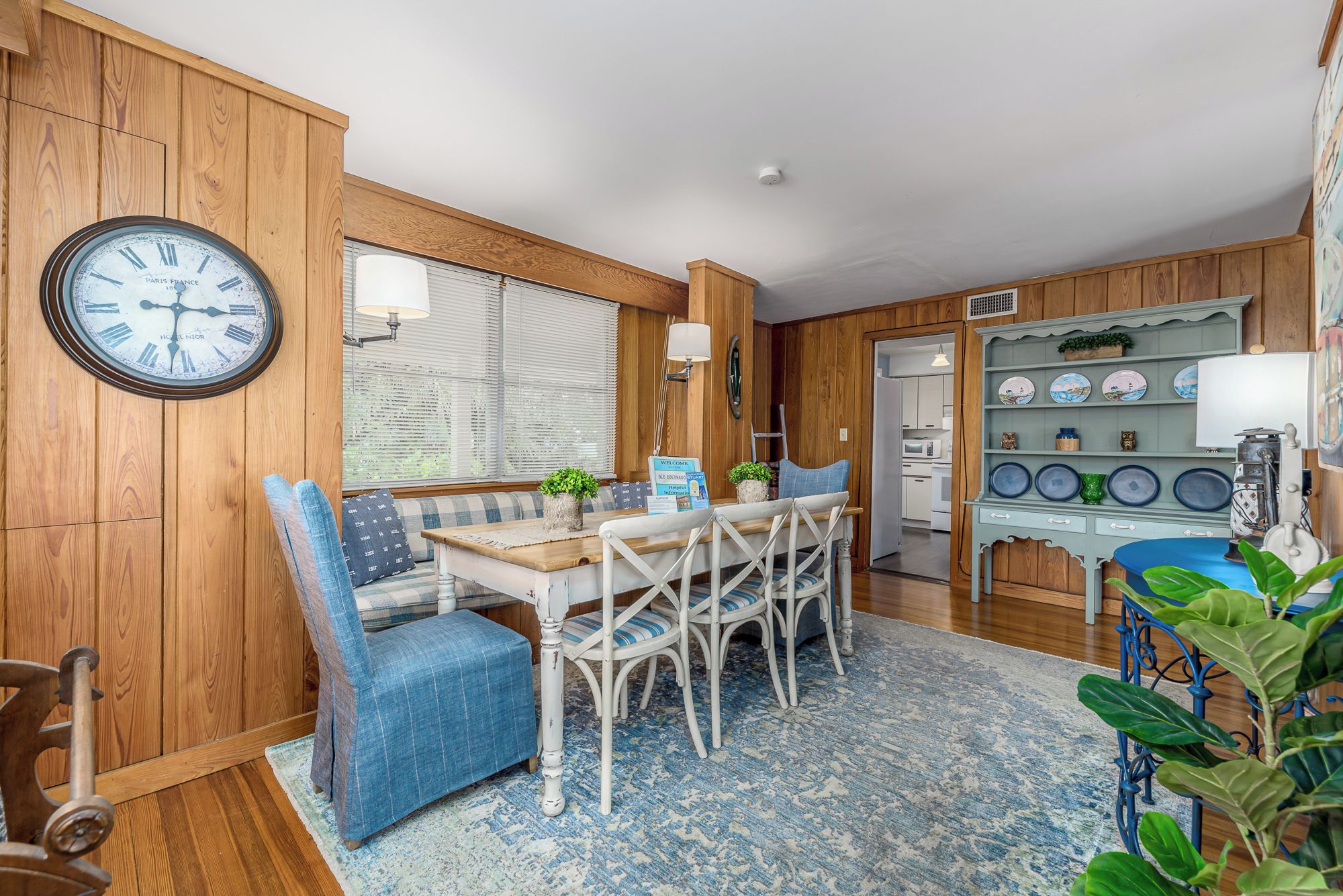 A cozy kitchen with wooden walls, a round clock, a dining table with chairs, and a shelf with plates.