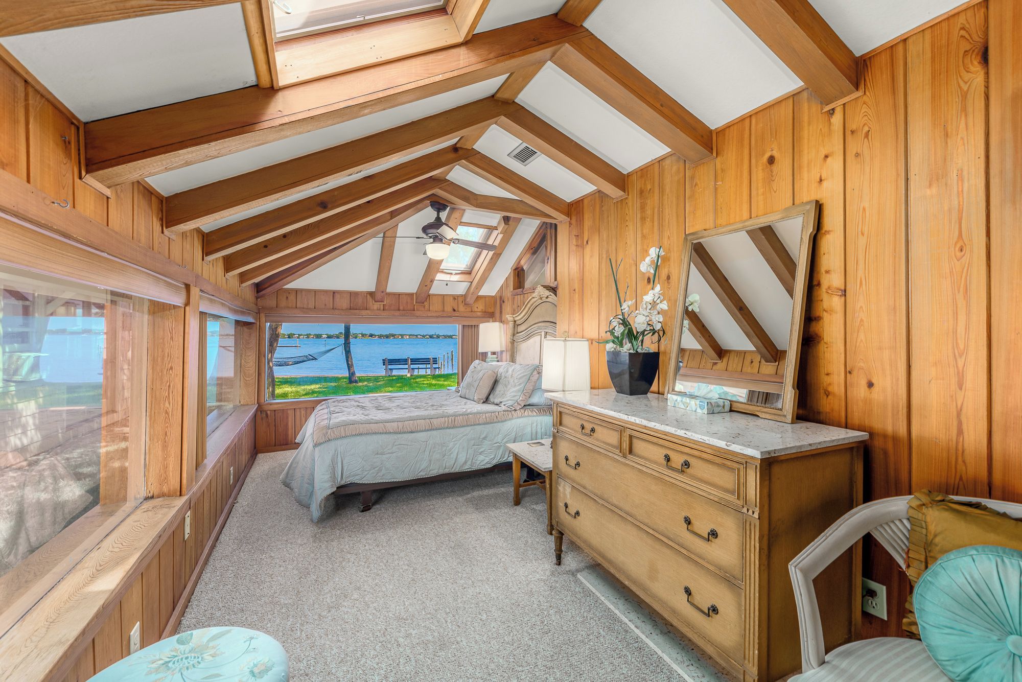 A bedroom with a bed, dresser, and a large mirror. There is a window with a view of a body of water and a dock.
