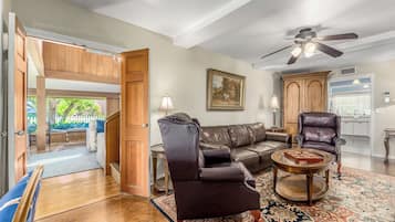 Un salon spacieux avec un canapé en cuir, un fauteuil en cuir et une table basse. Un tableau orne le mur, un ventilateur de plafond est installé au plafond, et une porte ouverte offre une vue sur l’extérieur.