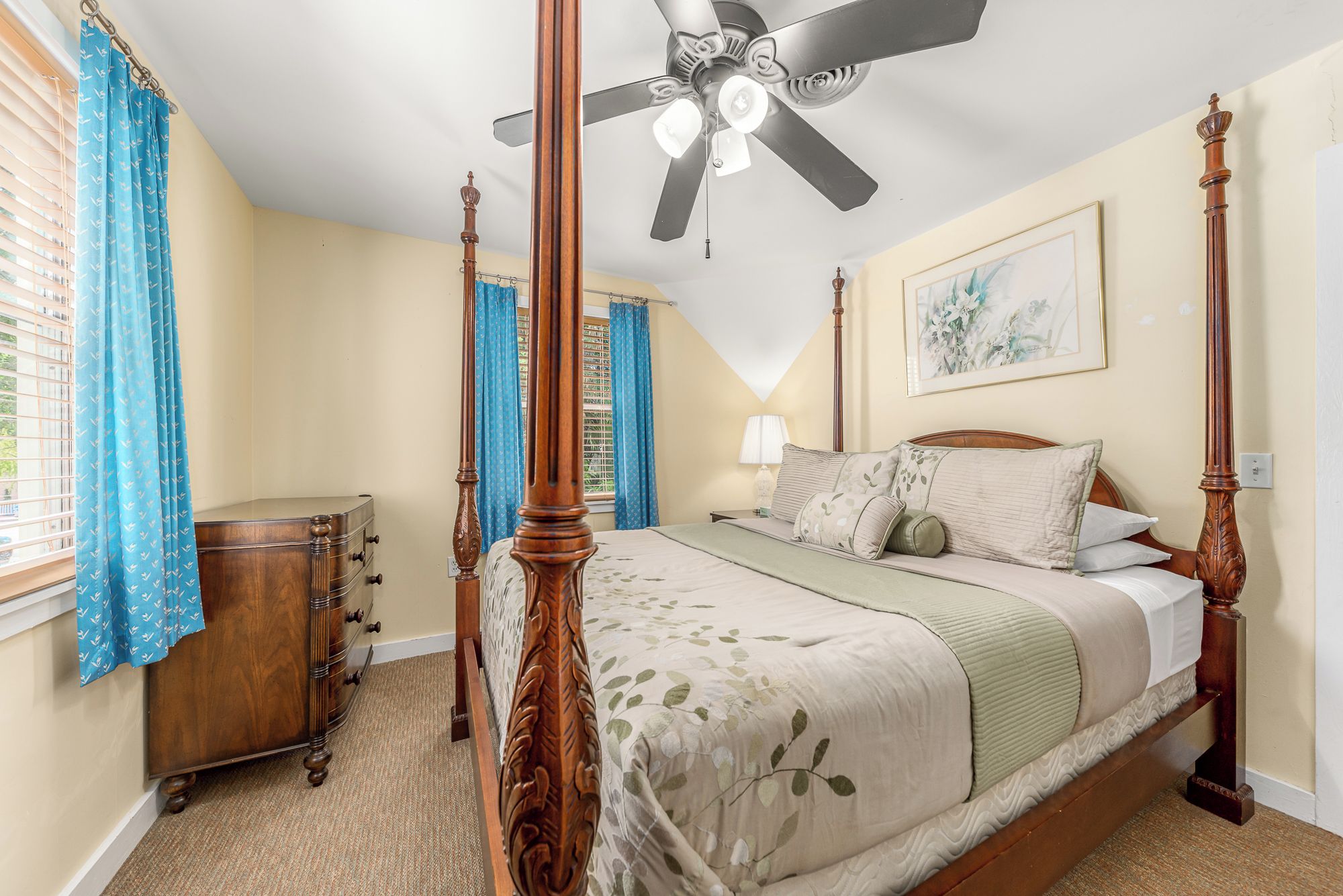 A four-poster bed with a floral bedspread, a ceiling fan, and a window with blue curtains.