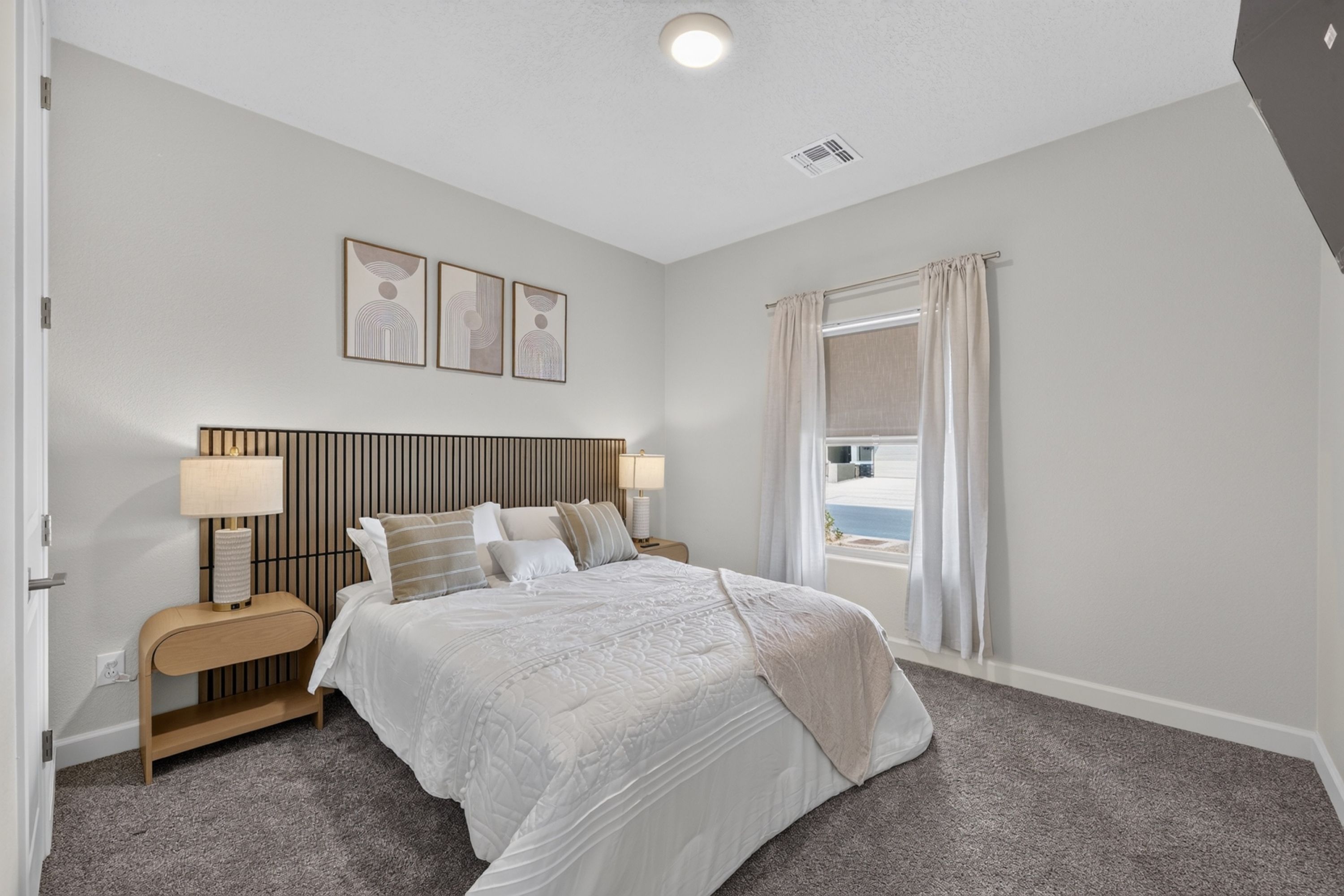 Quarto com cama, mesas de cabeceira, luminárias, janela com cortinas e quadros na parede.