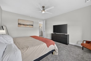 A hotel room with a bed, a flat-screen TV, a ceiling fan, a painting, and a kitchenette.
