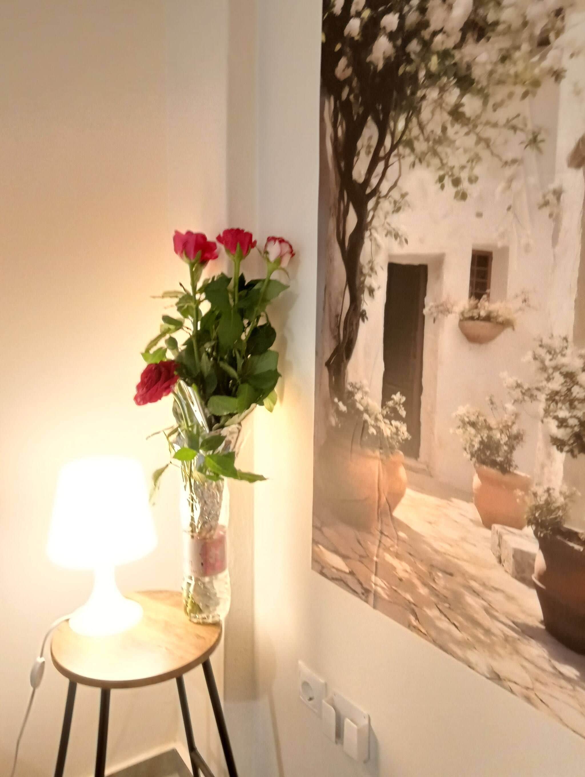 A room with a wall-mounted picture of a white building with a door and windows, a small table with a lamp, and a vase with flowers.