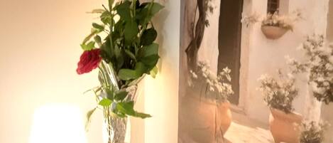 A room with a wall-mounted picture of a white building with a door and windows, a small table with a lamp, and a vase with flowers.