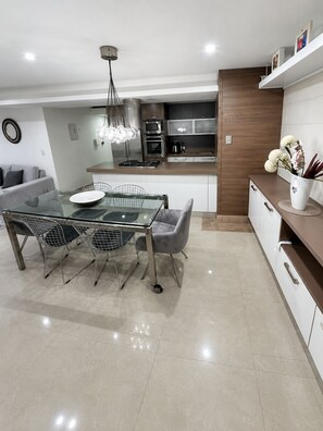 A modern kitchen with a dining area, featuring a glass table, metal chairs, and a pendant light.