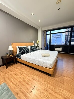 A modern hotel room with a large bed, a nightstand, a lamp, and a view of the city through large windows. - Cozy Studio Unit condo in the heart of Makati with a perfect city view. (Makati)