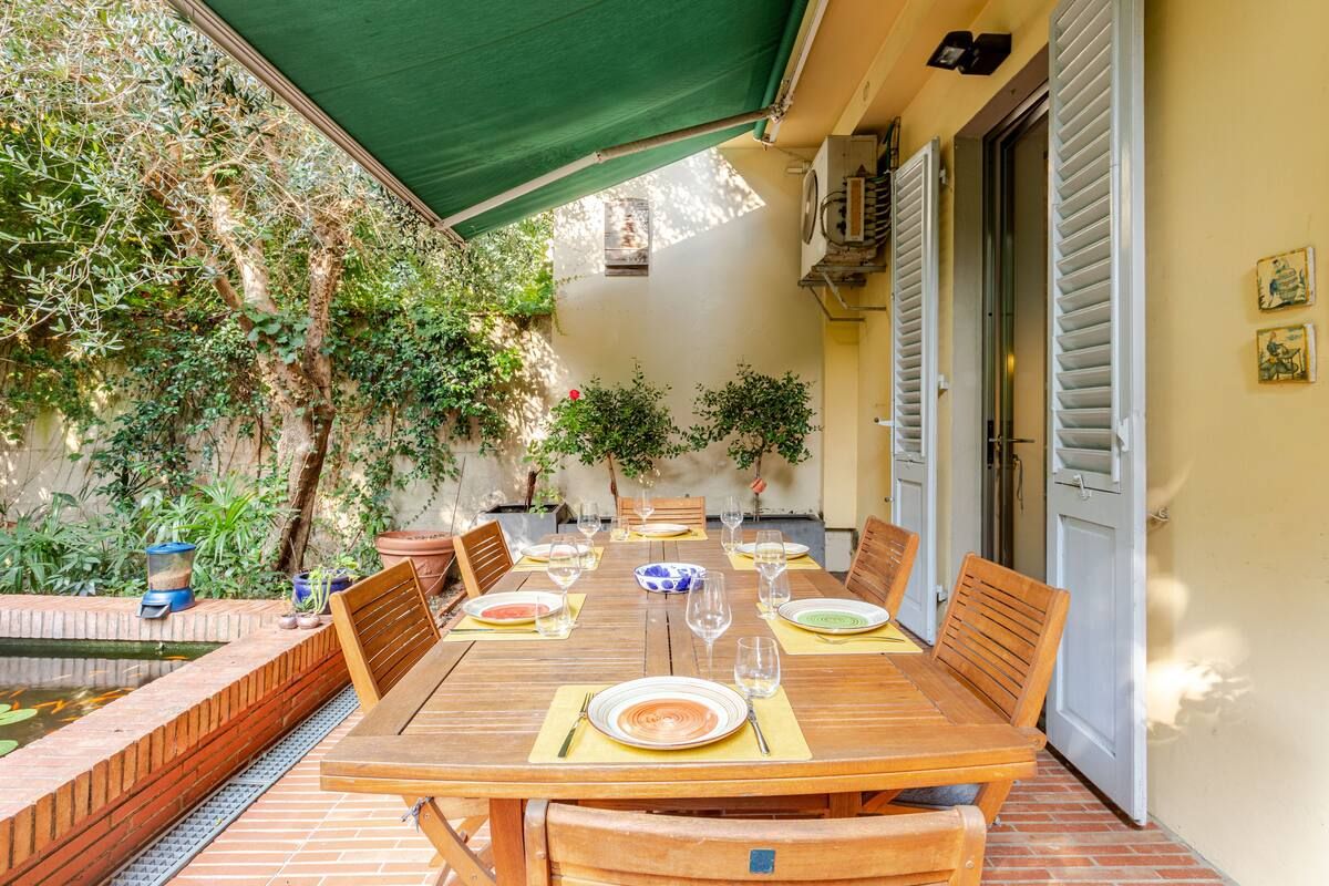 Zona pranzo all'aperto con tavolo e sedie in legno, circondata da piante e un muro in mattoni.