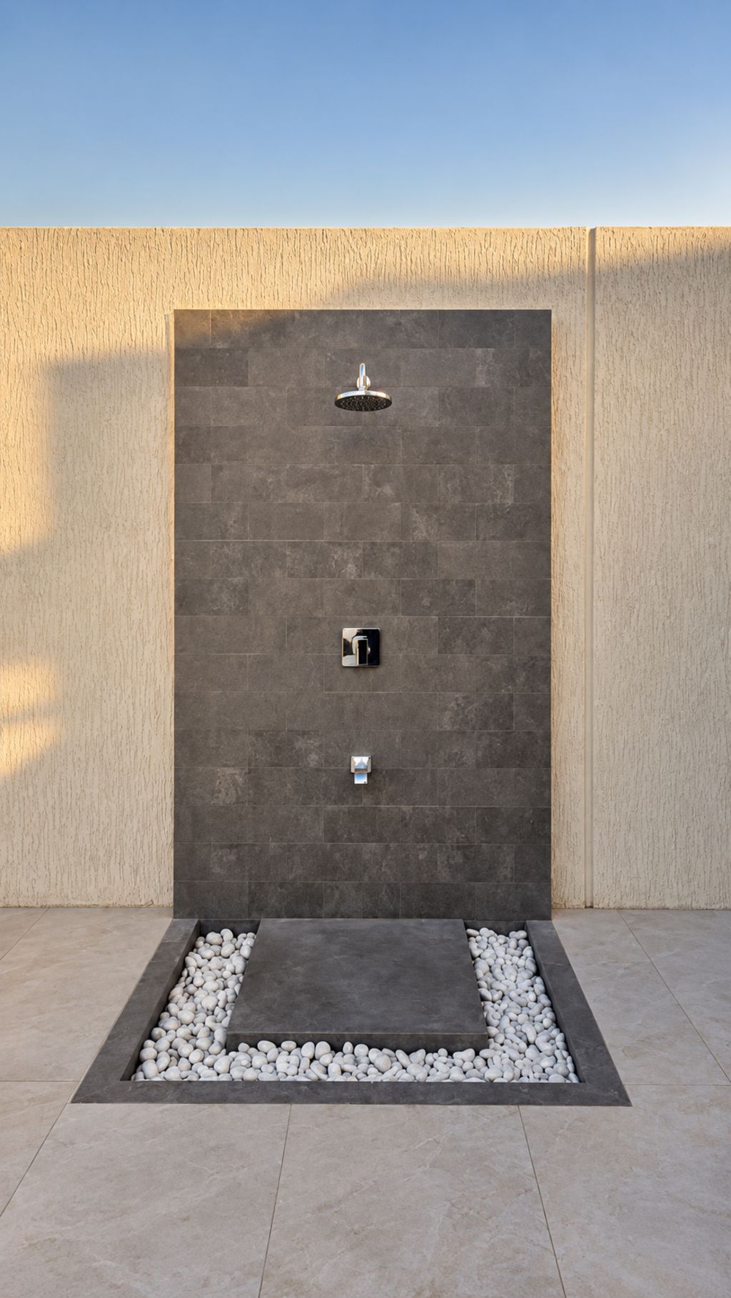 Eine moderne Außendusche mit einer dunkel gefliesten Wand, einer Bedienkonsole und einem Becken mit Kieselsteinen.