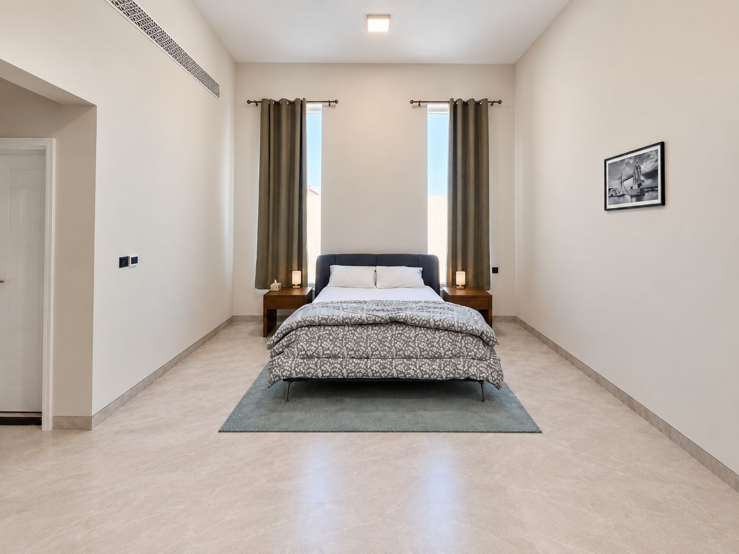 Ein Schlafzimmer mit einem zentrierten Bett, zwei Nachttischen mit Lampen, einer Tür links und einem gerahmten Bild an der Wand.