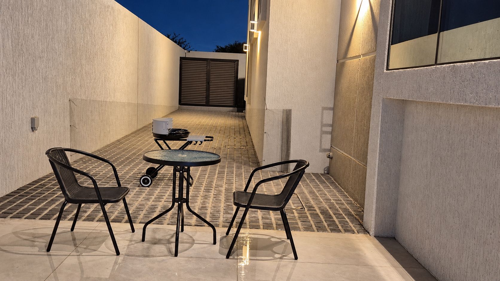 Een terras buiten met een tafel en twee stoelen, een barbecue en een gesloten poort op de achtergrond.