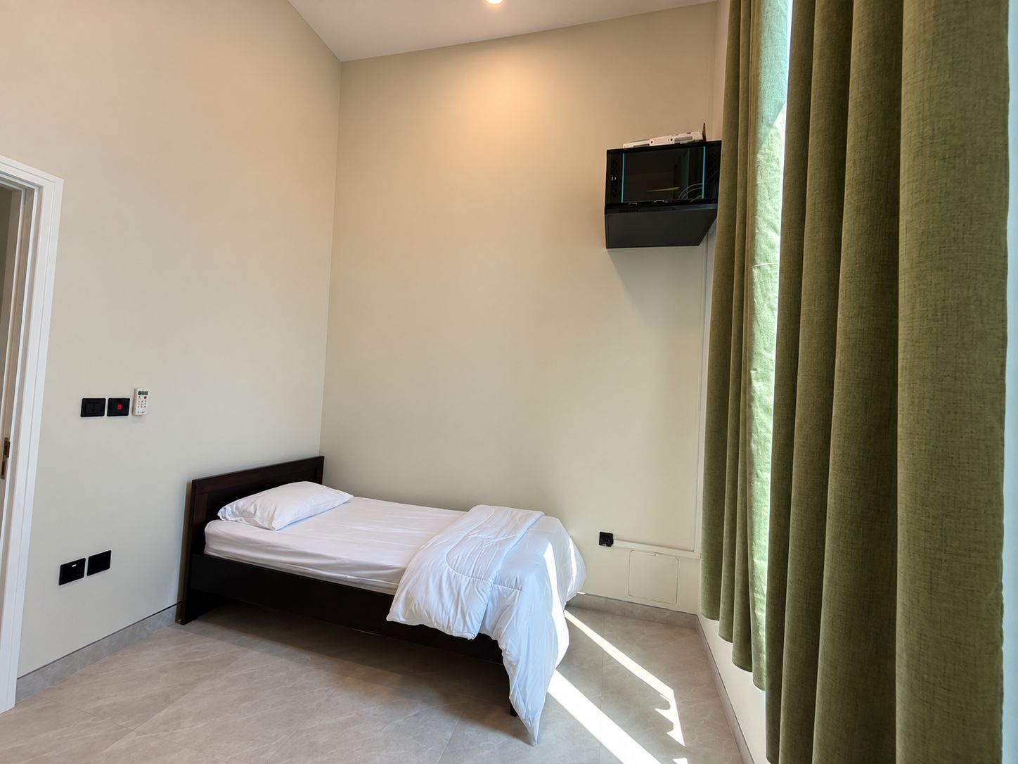 A single bed in a room with a wall-mounted TV and a door with a light switch.