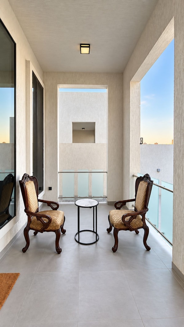 A balcony with two chairs and a small table, large windows, and a view of the sky.