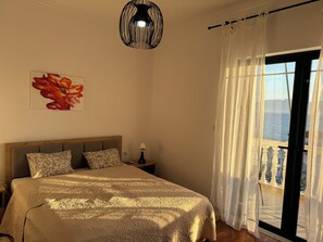 A bedroom with a bed, a nightstand, a lamp, a painting, and a view of the sea through a window with curtains. - Spacious 3 Bedroom Apartment with Stunning Sea View (Saranda)