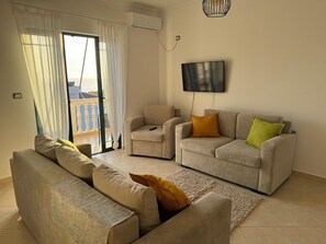 A living room with a sofa set, a television mounted on the wall, and a view through a glass door. - Spacious 3 Bedroom Apartment with Stunning Sea View (Saranda)