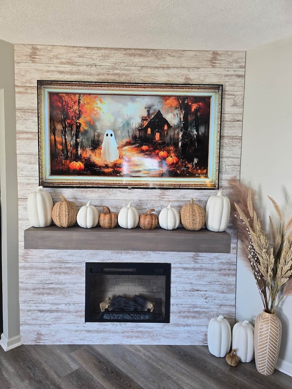 Una chimenea con un fuste rústico, un cuadro enmarcado encima y calabazas decorativas sobre el fuste.