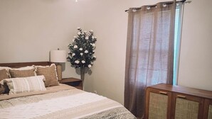 Un dormitorio con cama, mesita de noche, lámpara, florero con flores, ventana con cortinas y ventilador de techo.