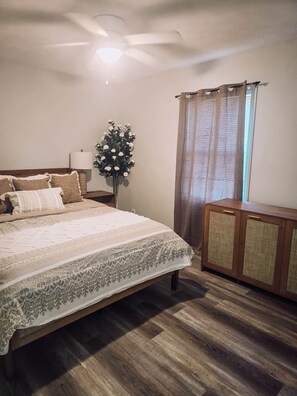 Un dormitorio con cama, mesita de noche, lámpara, florero con flores, ventana con cortinas y ventilador de techo.