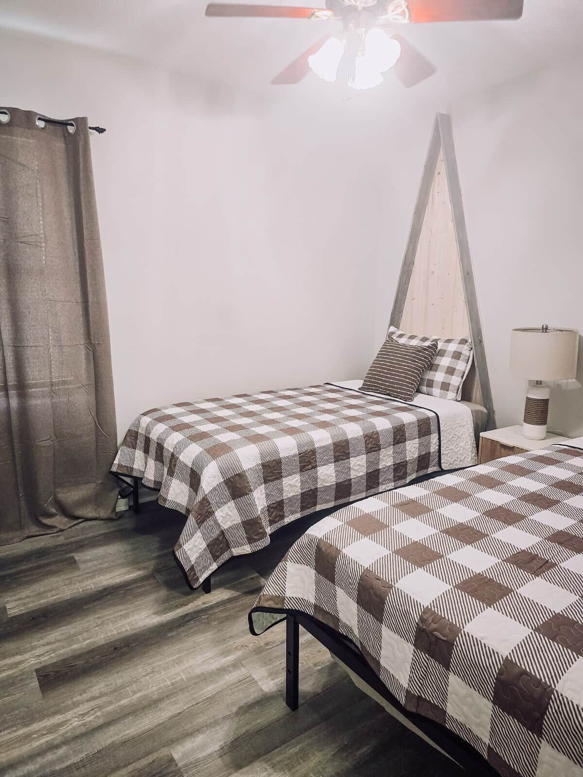 A room with two beds, a ceiling fan, a nightstand with a lamp, and a window with curtains.