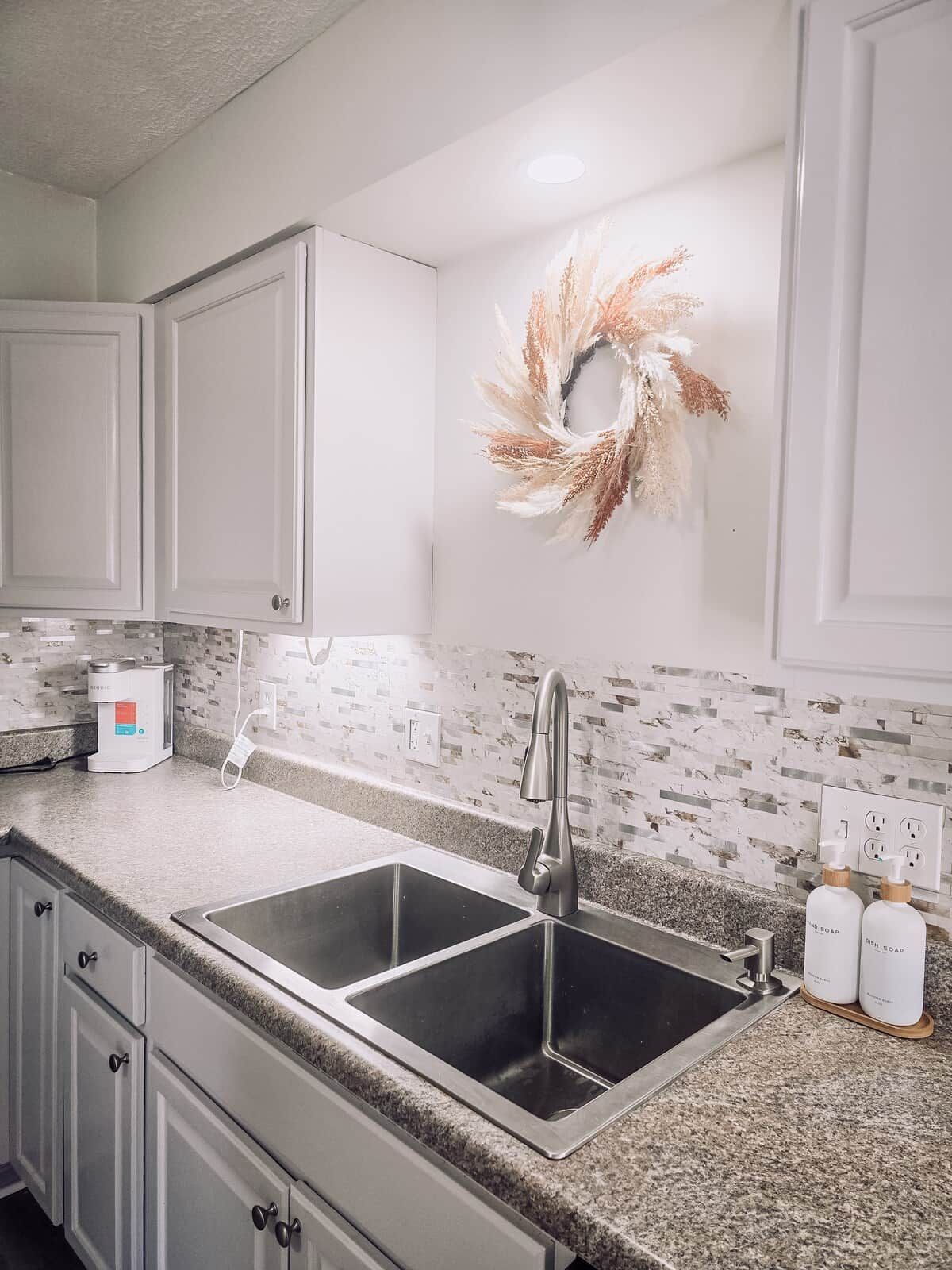 Una cocina con dos lavamanos de acero inoxidable, un protector contra salpicaduras blanco y un adorno decorativo en la pared.