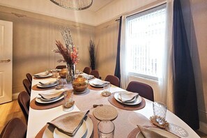 A dining room with a long table set for a meal, featuring a vase with dried flowers, a textured centerpiece, and a view of the city through large windows. - 5-Bed Home | 10 Mins to Etihad & City Centre (Greater Manchester)
