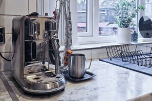 Eine Küche mit einer Edelstahl-Espressomaschine, einer Metalltasse und einem Fenster mit Blick auf Gebäude.