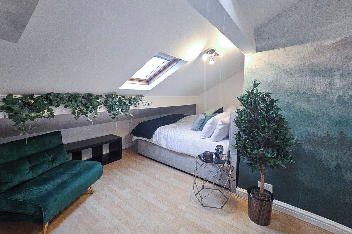 Une chambre à coucher aménagée dans un espace de type « loft », avec un puits de lumière, un canapé vert, un lit avec des coussins, une petite table et une plante en pot.