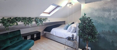 A loft bedroom with a skylight, a green sofa, a bed with pillows, a small table, and a potted plant.