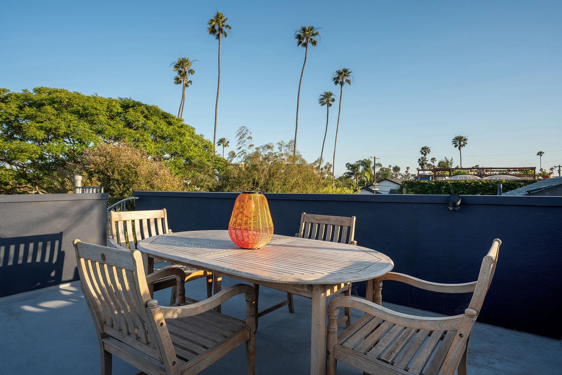 An outdoor patio with a round table and four wooden chairs, surrounded by a dark wall and palm trees in the background.