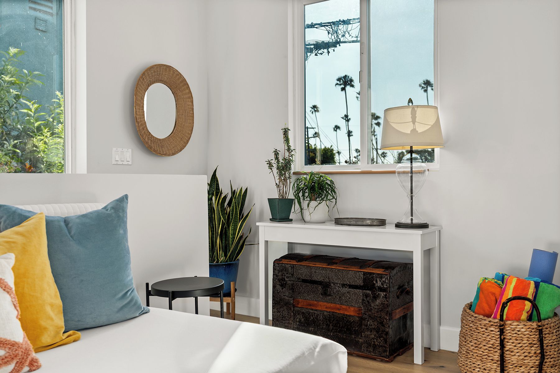 Een slaapkamer met een bed, een ronde spiegel, een raam met uitzicht op palmbomen, een lamp en een klein tafeltje met planten.