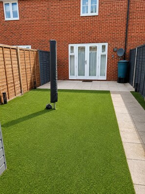 A backyard with artificial grass, a wooden fence, a black pole, and a brick house with windows and a door. - Modern 3-Bed Home Free Driveway Parking, Fast Wi-Fi (Ebbsfleet Valley)