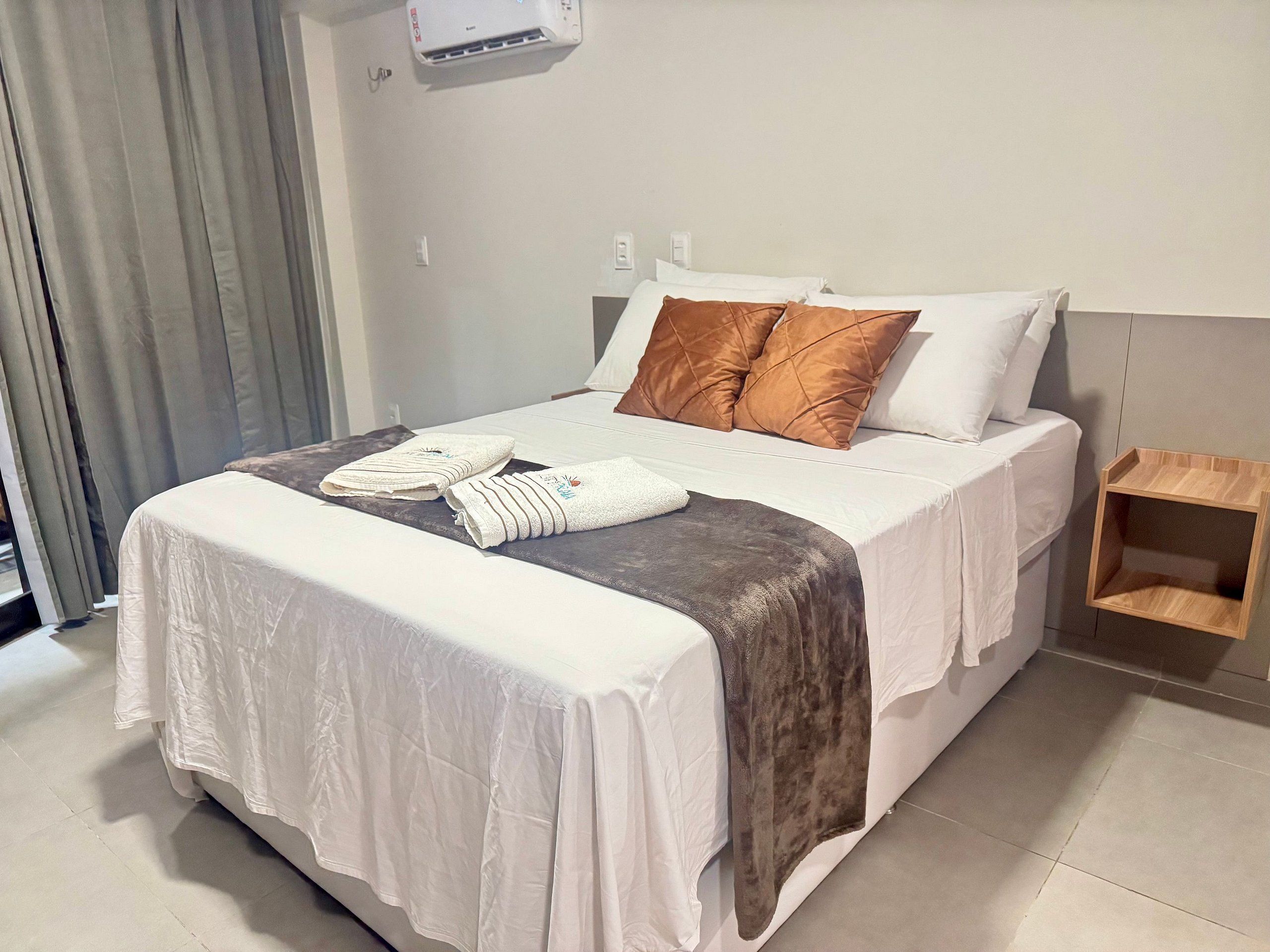Una cama bien hecha con ropa de cama blanca y marrón, una mesita de noche pequeña y un aire acondicionado de pared.