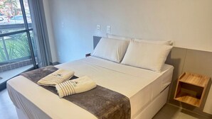 A hotel room with a bed, a window, and a small wooden side table. - Season Smart Flats By Flat na Praia (Ipojuca)