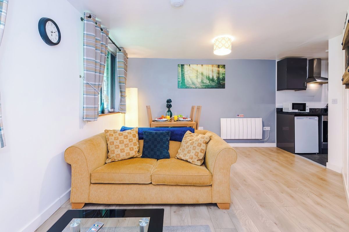 A living room with a sofa, a dining table with chairs, a kitchen area with appliances, and a clock on the wall.