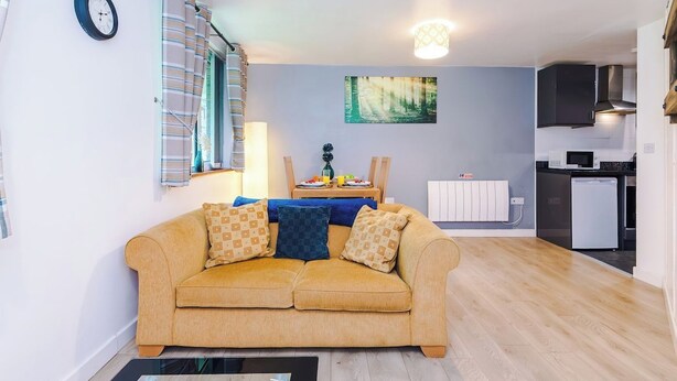 A living room with a sofa, a dining table with chairs, a kitchen area with appliances, and a clock on the wall. - Cosy 1-Bed Apartment Wigan | Free Parking (Greater Manchester)