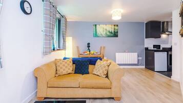 A living room with a sofa, a dining table with chairs, a kitchen area with appliances, and a clock on the wall.