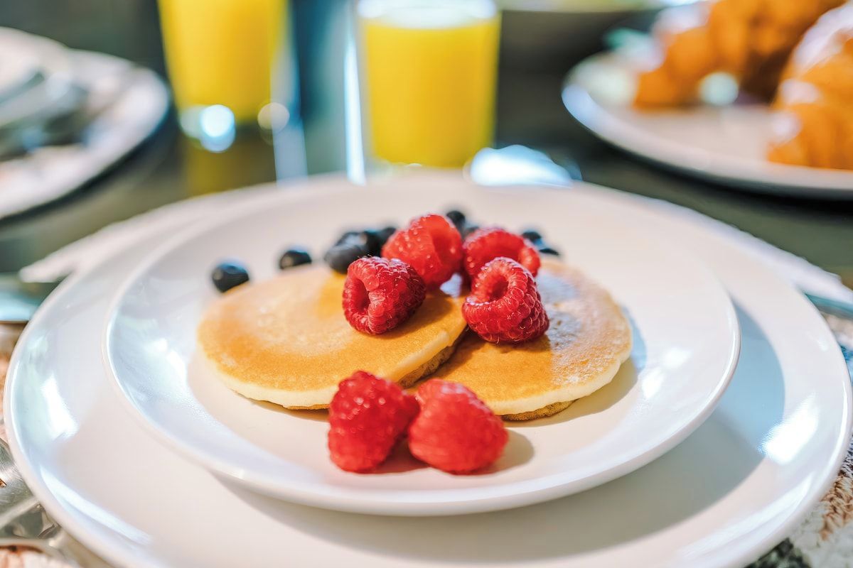 En morgenmadstallerken med pandekager, toppet med hindbær og blåbær, et glas appelsinjuice og en croissant ved siden af.
