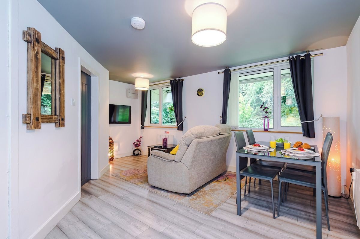 A modern living room with a sofa, dining table, and chairs. There is a TV mounted on the wall, and a window with curtains.