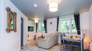 Ein modernes Wohnzimmer mit einer Couch, einem Esstisch und Stühlen. Ein Fernseher ist an der Wand montiert, und ein Fenster mit Vorhängen ist vorhanden.
