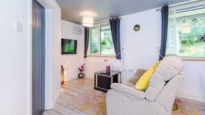 Ein modernes Wohnzimmer mit einer Couch, einem kleinen Tisch und einem Fernseher. Es gibt ein Fenster mit Vorhängen, eine Tür und eine Deckenleuchte.