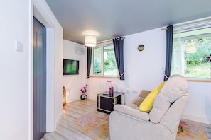 A modern living room with a sofa, a small table, and a TV. There is a window with curtains, a door, and a light fixture. - Stylish 1-Bed Apartment in Wigan | Free Parking (Greater Manchester)