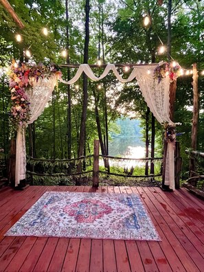 An outdoor wooden deck with a patterned rug, surrounded by a wooden fence and draped with white fabric and string lights.
