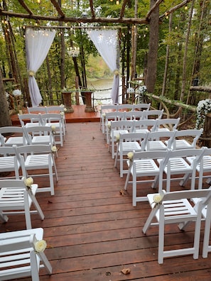 A wooden deck with white chairs arranged for an event, surrounded by trees and a body of water.