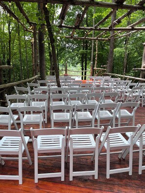 An outdoor seating area with white chairs arranged in rows, surrounded by trees and a wooden deck.
