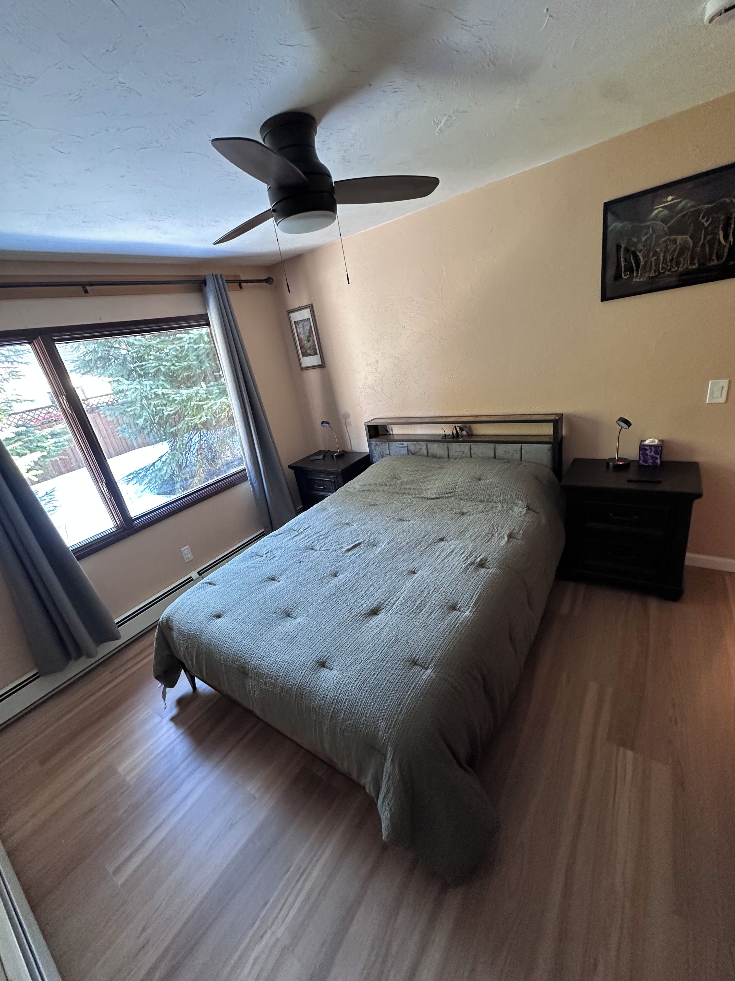 Un dormitorio con cama, ventilador de techo, mesita de noche, lámpara y ventana con cortinas.