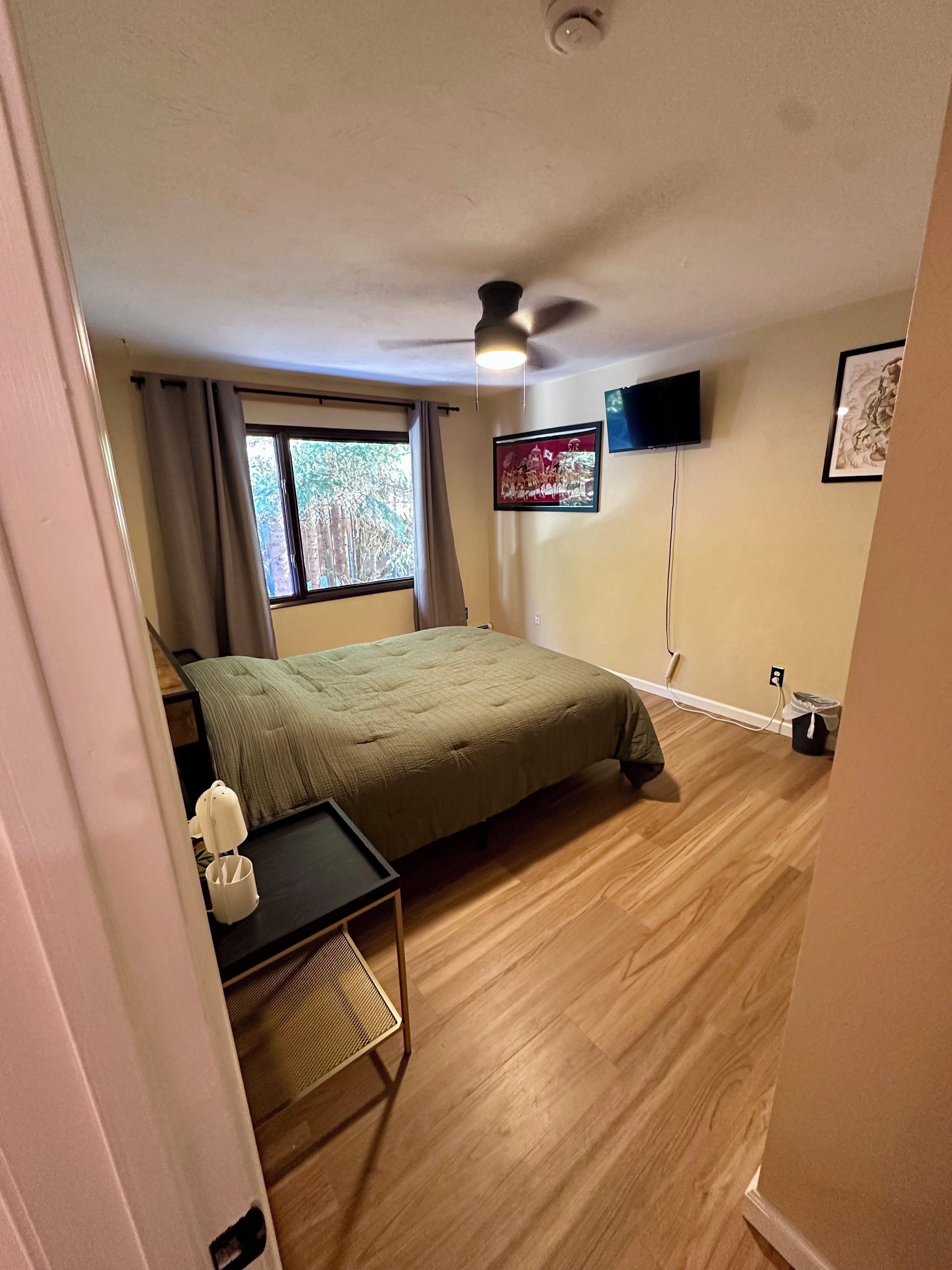 A bedroom with a bed, a desk, a chair, a ceiling fan, a window with curtains, and a wall-mounted TV.