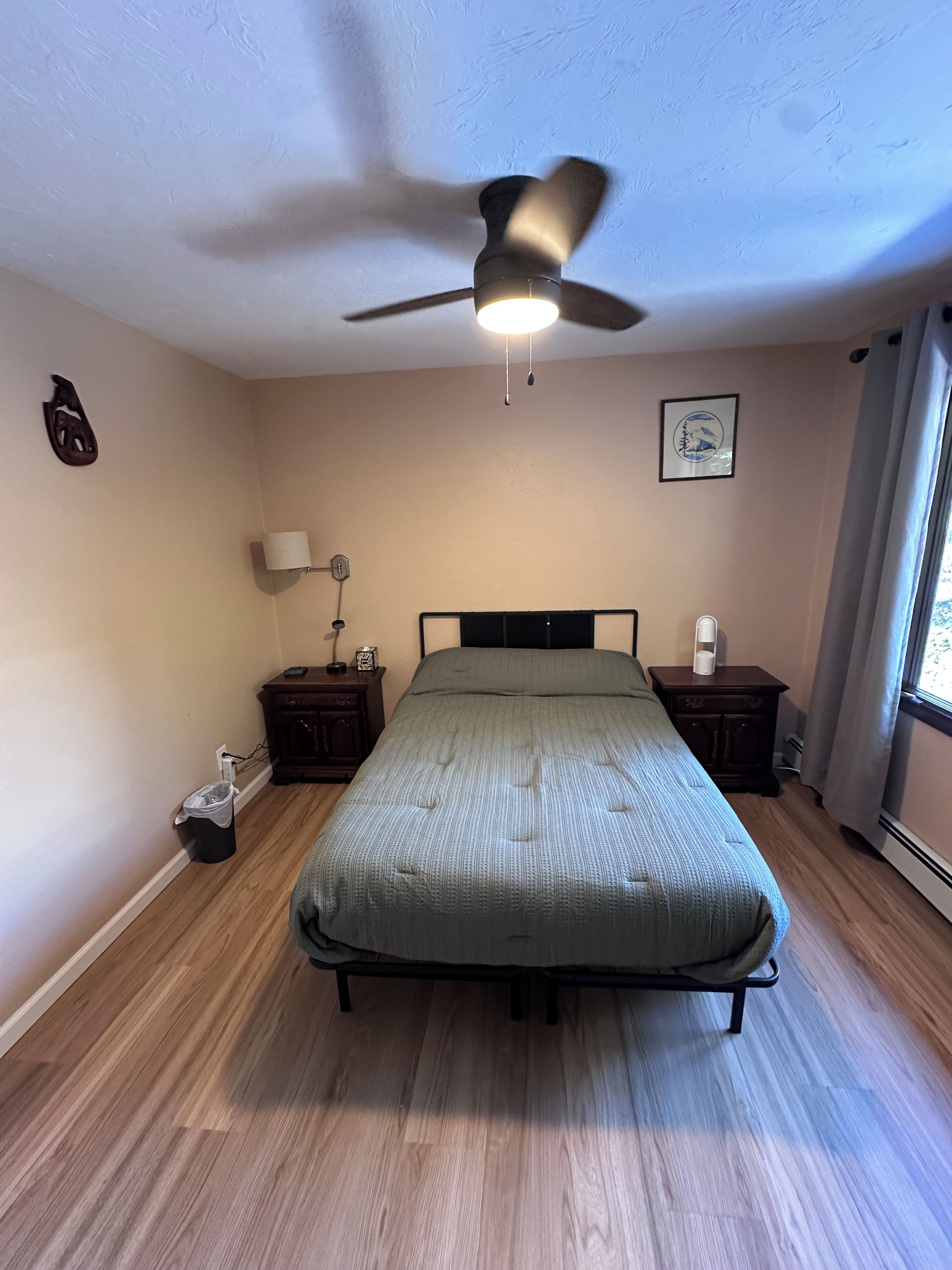 Une chambre à coucher avec un lit, un ventilateur de plafond, une table de chevet avec une lampe et une fenêtre avec des rideaux.
